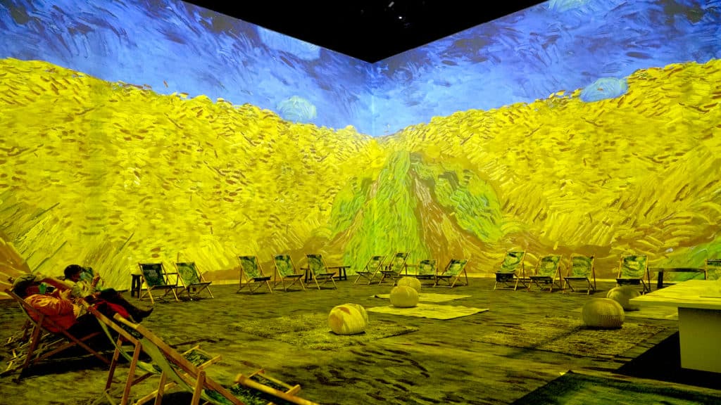 people sit on deck chairs in a room where Van Gogh's wheat fields painting is projected on the wall