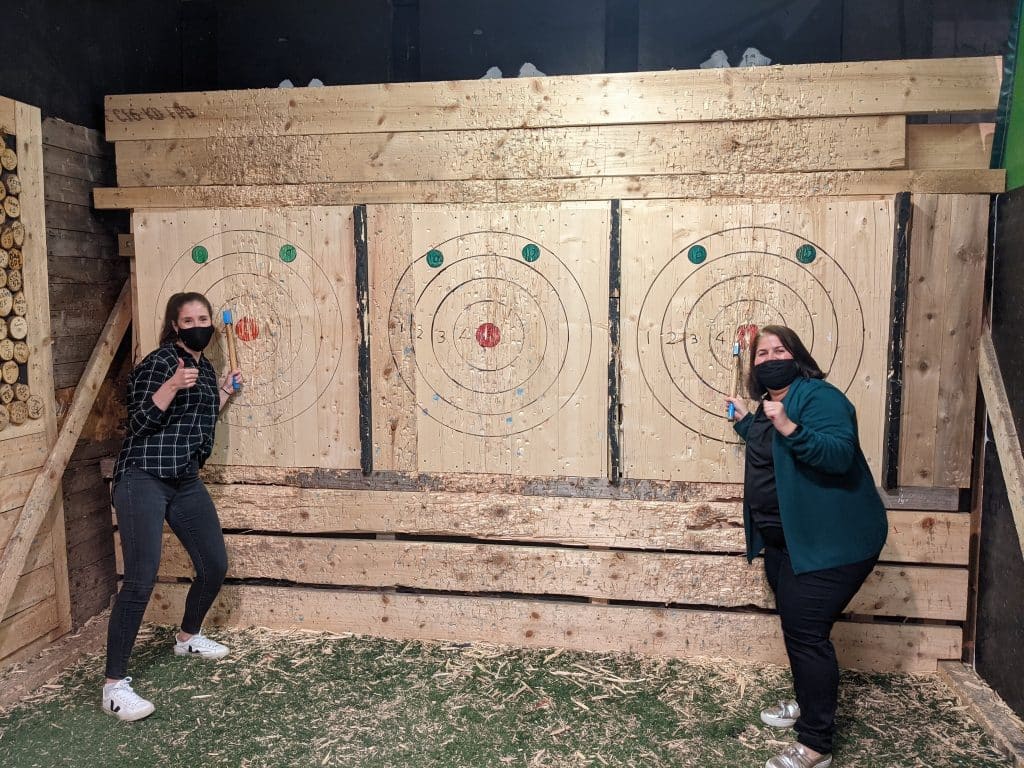 Two women give the thumbs up to the camera, holding the axes they've just thrown at a target.