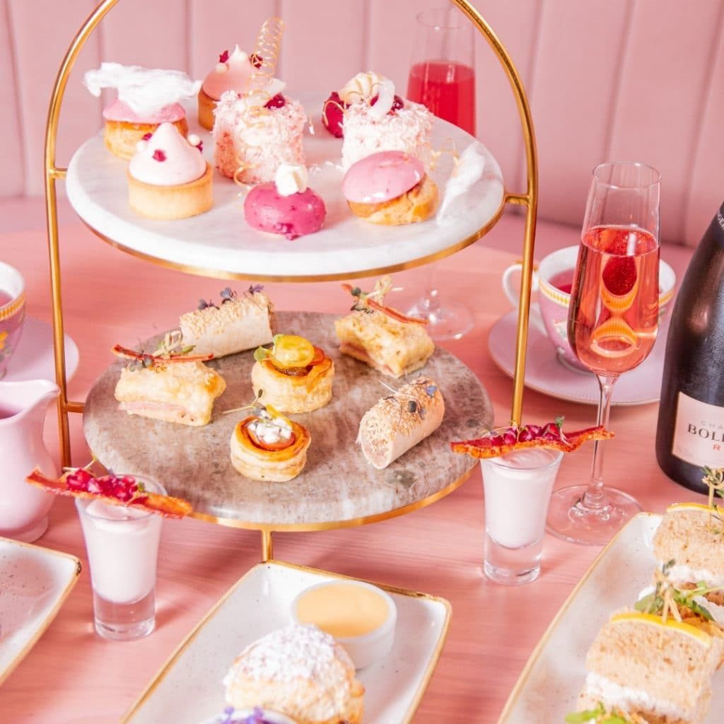 Pink, iced mini desserts and sandwiches with pink champagne on a pink table.