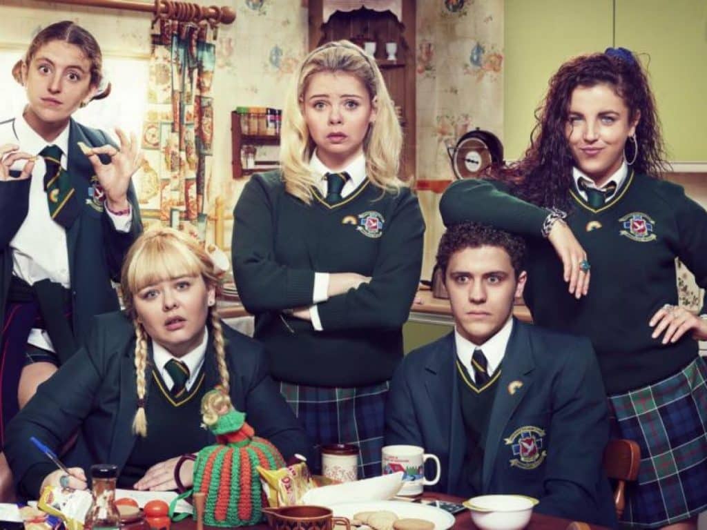 The Derry Girls stand and sit around a table in their school uniforms.