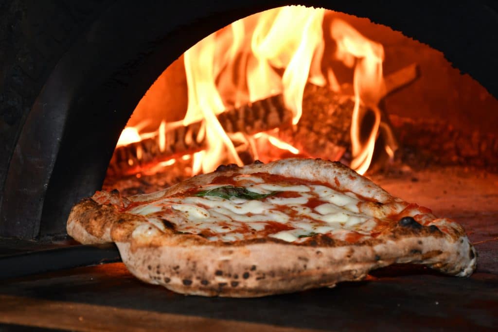 A close-up image of a cheesy pizza being put into a woodburning oven.