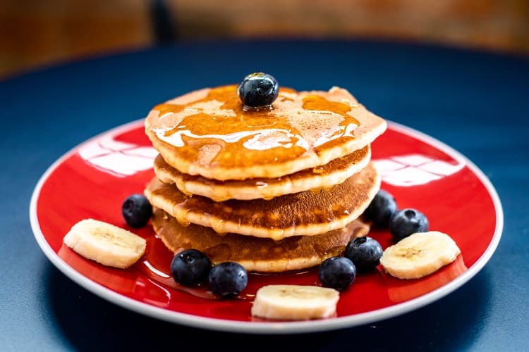 A stack of American pancakes, topped woth syrup, bananas and blueberries.