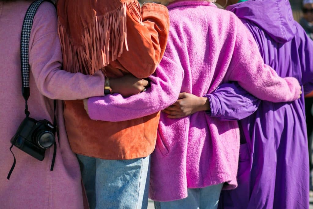 Four women in purple, pink and orange coats stand arm in arm. 